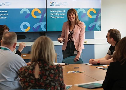Woman standing speaking to a group of four people seated around a table. Behind her are two display boards with graphics and text that reads Management Development Programme